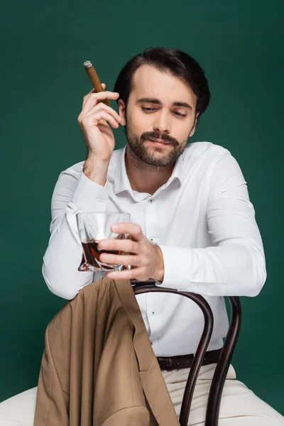 Man with mustache holding cigar and glass of brandy while sitting on chair on dark green — Stock Photo