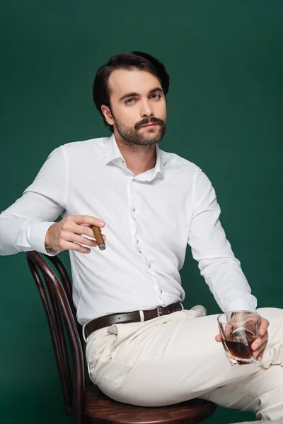 Man in white shirt holding cigar and glass of whiskey while sitting on chair on dark green — Stock Photo