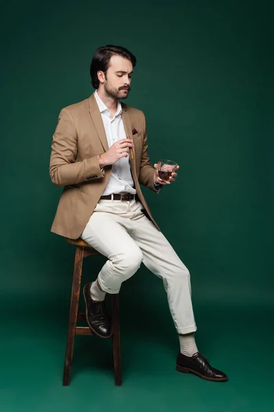 Longitud completa del hombre en blazer celebración de cigarro y vaso de whisky mientras está sentado en la silla en verde oscuro - foto de stock