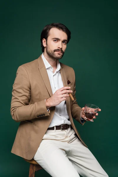 Man in blazer holding cigar and glass of whiskey while sitting on chair on dark green — Stock Photo