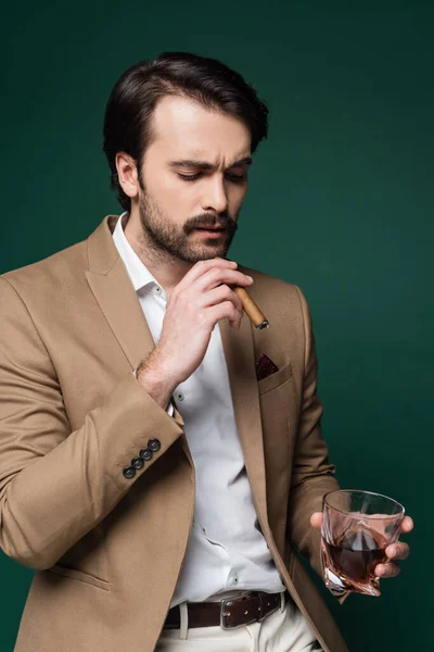 Man in blazer holding cigar and glass of scotch on dark green — Stock Photo