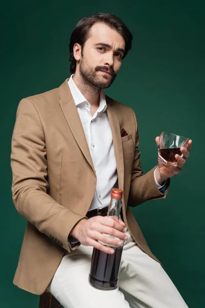 Mann im Blazer hält Flasche und Glas Scotch auf dunkelgrün — Stockfoto