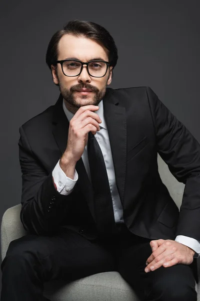 Retrato de empresário de terno e óculos sentado na poltrona e olhando para a câmera no cinza escuro — Fotografia de Stock