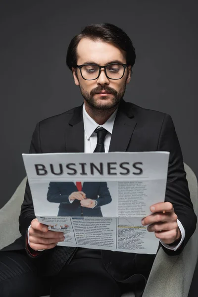 Homme d'affaires en costume et lunettes assis sur un fauteuil et lisant un journal d'affaires sur gris foncé — Photo de stock