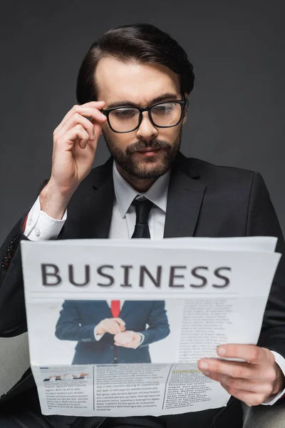 Businessman adjusting glasses while sitting on armchair and reading business newspaper on dark grey — Stock Photo