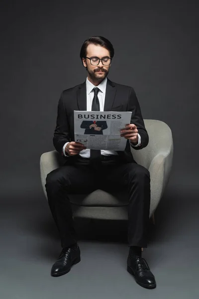Longitud completa de hombre de negocios en traje y gafas sentado en el sillón y leyendo el periódico de negocios en gris oscuro - foto de stock
