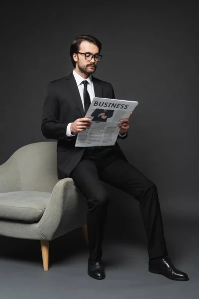 Full length of businessman in suit and glasses leaning on armchair and reading business newspaper on dark grey — Stock Photo