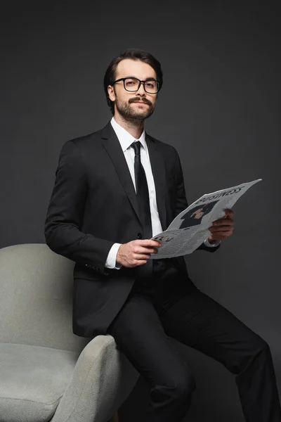 Бізнесмен в костюмі і окулярах спирається на крісло і тримає ділову газету на темно-сірому — стокове фото