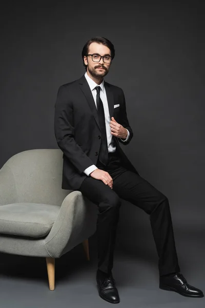 Businessman in suit and glasses leaning on armchair on dark grey — Stock Photo