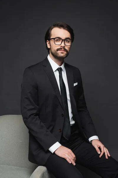 Businessman in formal wear and glasses leaning on armchair on dark grey — Stock Photo