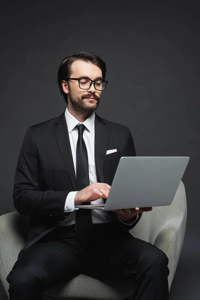 Uomo d'affari in abito e occhiali seduto sulla poltrona e utilizzando il computer portatile su grigio scuro — Foto stock