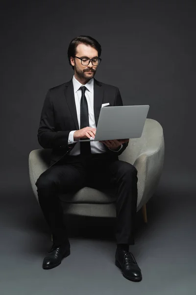 Toute la longueur de l'homme d'affaires en costume et lunettes assis sur un fauteuil et en utilisant un ordinateur portable sur gris foncé — Photo de stock