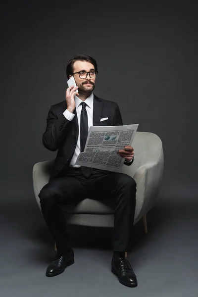 Homme d'affaires en costume et lunettes assis sur un fauteuil et parlant sur smartphone tout en tenant journal sur gris foncé — Photo de stock