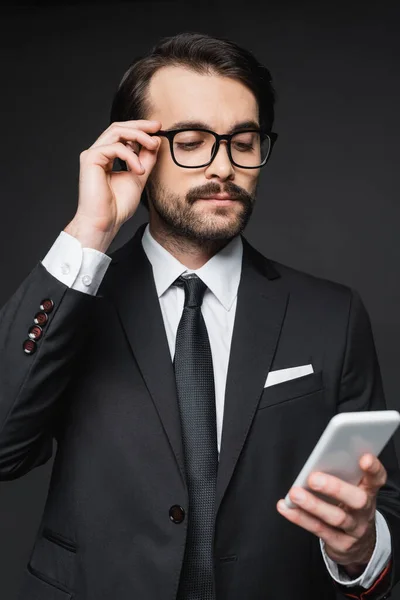 Uomo d'affari in giacca e cravatta e occhiali che guarda smartphone su grigio scuro — Foto stock