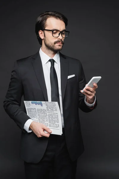 Homme d'affaires en costume et lunettes tenant journal et utilisant un smartphone sur gris foncé — Photo de stock