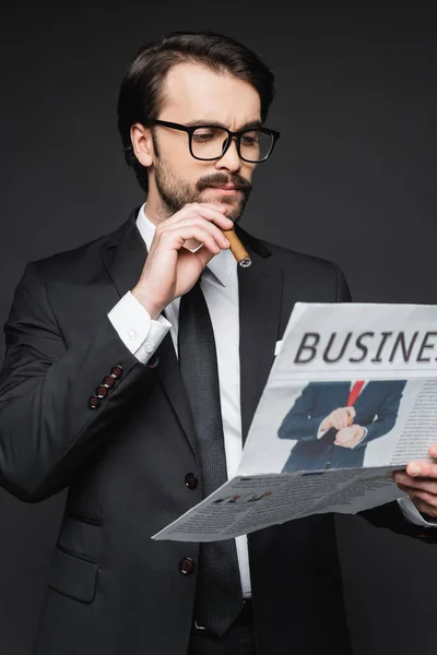 Hombre de traje y gafas leyendo periódico de negocios y sosteniendo cigarro en gris oscuro - foto de stock