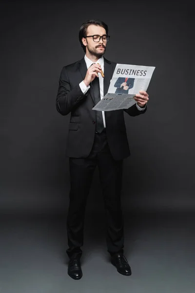 Full length of man in suit and glasses reading business newspaper and holding cigar on dark grey — Stock Photo