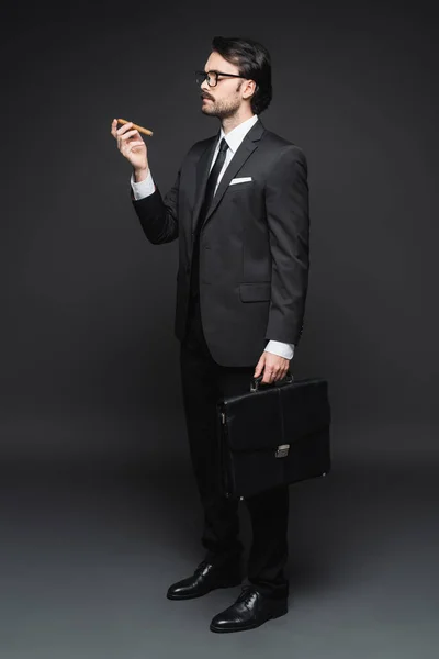 Повнометражний чоловік у костюмі та окулярах тримає сигару та шкіряний портфель на темно-сірому — стокове фото