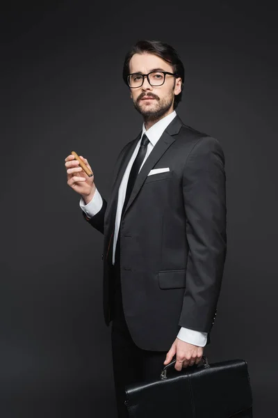Businessman in suit and glasses holding cigar and leather briefcase on dark grey — Stock Photo