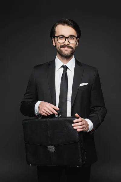 Homme d'affaires souriant en costume et lunettes tenant une mallette en cuir sur gris foncé — Photo de stock