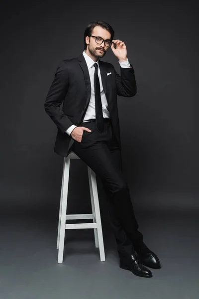 Full length of businessman adjusting glasses and leaning on chair while posing with hand in pocket on dark grey — Stock Photo