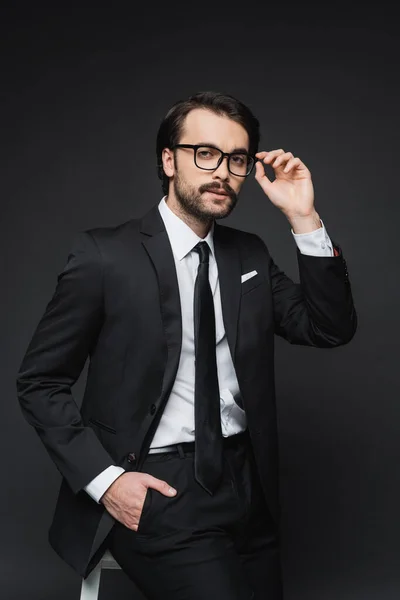 Бізнесмен регулює окуляри і спирається на стілець, позуючи рукою в кишені на темно-сірому — стокове фото