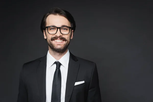 Cheerful businessman with mustache in glasses smiling on dark grey — Stock Photo
