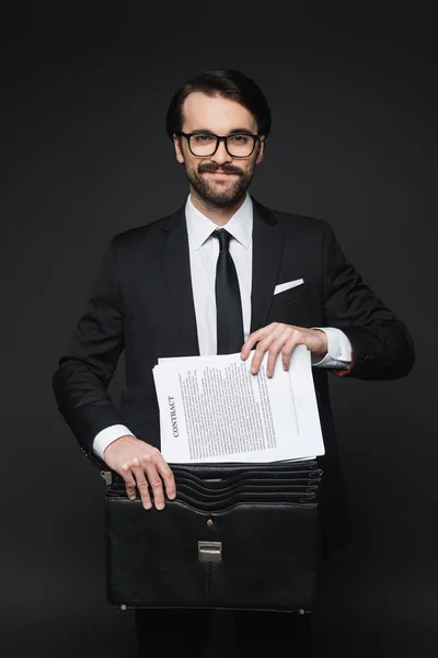 Alegre hombre de negocios con bigote en gafas poniendo contrato en maletín de cuero en gris oscuro - foto de stock