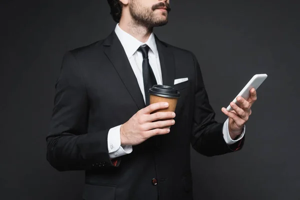 Vista recortada de hombre de negocios con bigote sosteniendo taza de papel y teléfono inteligente en gris oscuro — Stock Photo