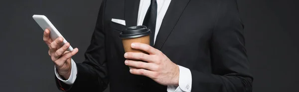 Vista recortada de hombre de negocios sosteniendo taza de papel y teléfono inteligente en gris oscuro, bandera - foto de stock