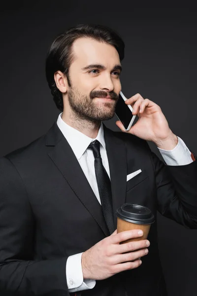 Homme d'affaires souriant avec moustache tenant une tasse en papier et parlant sur smartphone sur gris foncé — Photo de stock