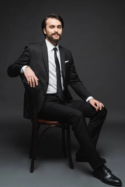 Full length of smiling businessman with mustache sitting on wooden chair on dark grey — Stock Photo