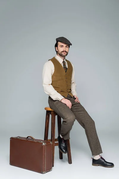 Hombre con bigote y ropa de estilo retro sentado en silla cerca del bolso de viaje aislado en gris - foto de stock