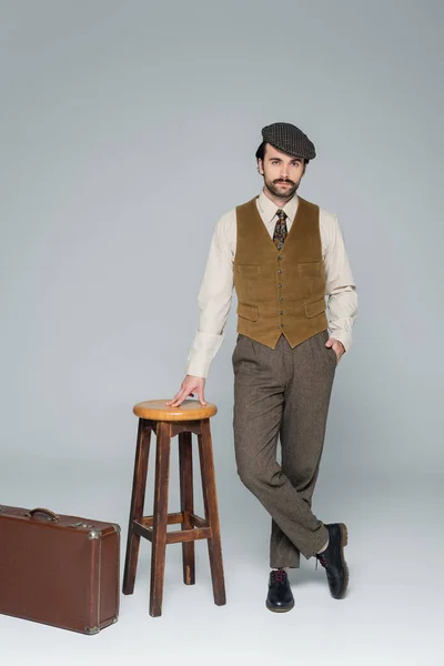 Full length of man with mustache and retro style clothing standing with hand in pocket near luggage and chair on grey — Stock Photo