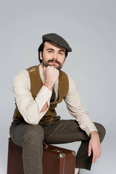 Mann mit Schnurrbart und Kleidung im Retro-Stil sitzt auf Reisetasche isoliert auf grau — Stockfoto