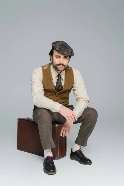 Full length of man with mustache and retro style clothing sitting on travel bag on grey — Stock Photo