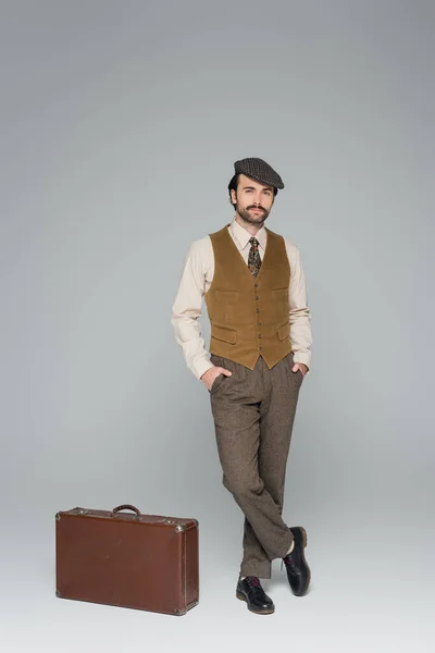 Full length of man with mustache and retro style clothing standing with hands in pockets near travel bag on grey — Stock Photo