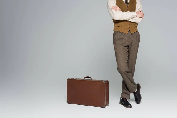 Cropped view of man in retro style clothing standing with crossed arms near travel bag on grey — Stock Photo