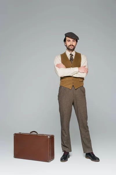 Full length of man in retro style clothing standing with crossed arms near travel bag on grey — Stock Photo