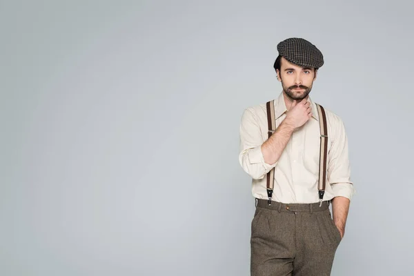 Пенсіонер у сорочці з підтяжками та капелюхом ретро стилю позує з рукою в кишені ізольовано на сірому — стокове фото