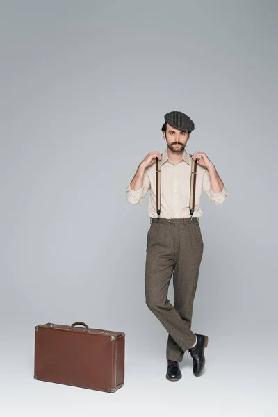 Longitud completa del hombre en ropa de estilo retro y sombrero de pie con las piernas cruzadas cerca de la bolsa de viaje en gris - foto de stock