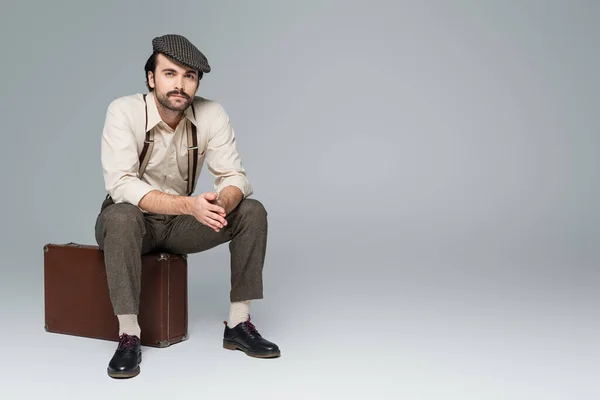 Full length of man with mustache in retro style clothing and hat sitting on travel bag on grey — Stock Photo