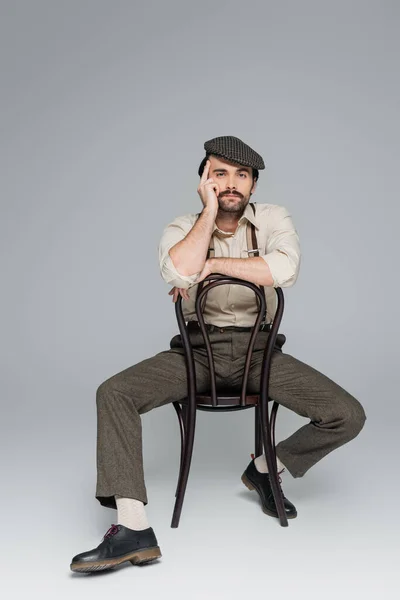 Full length of man with mustache in retro style clothing and hat sitting on wooden chair on grey — Stock Photo