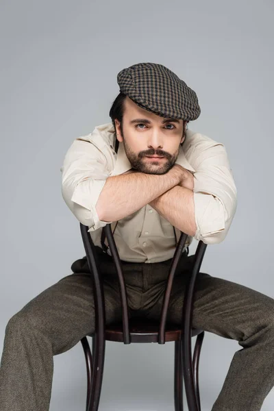 Homme avec moustache dans des vêtements de style rétro et chapeau assis sur une chaise en bois sur gris — Photo de stock