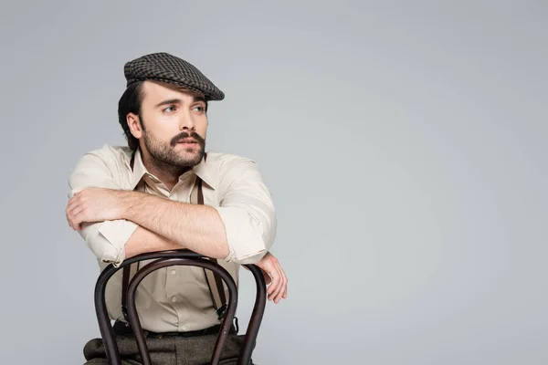 Homme avec moustache dans des vêtements de style rétro et chapeau assis sur une chaise en bois sur gris — Photo de stock