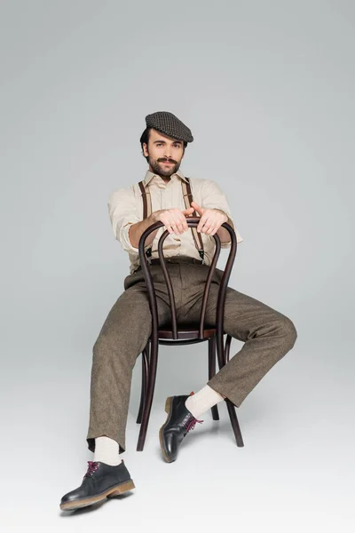 Pleine longueur de l'homme avec moustache dans des vêtements de style rétro et chapeau assis sur une chaise en bois sur gris — Photo de stock