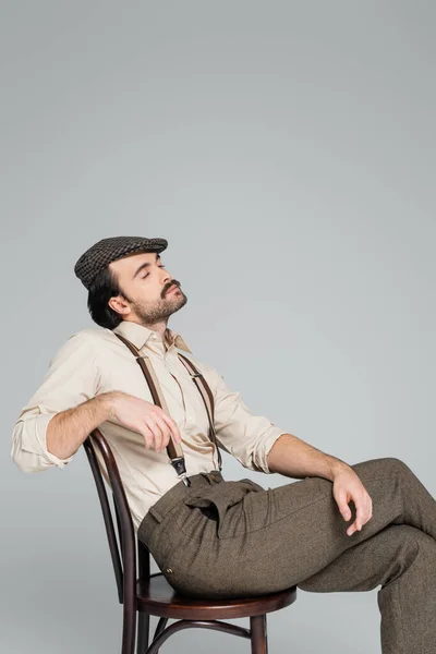 Homme dans des vêtements de style rétro et les yeux fermés assis sur une chaise en bois sur gris — Photo de stock