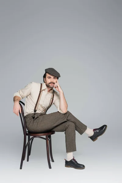 Longitud completa del hombre con bigote en ropa de estilo vintage y sombrero sentado en silla de madera en gris — Stock Photo