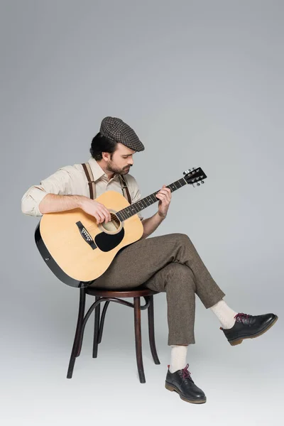 Мужчина в полный рост с мусташкой в одежде в стиле ретро и шляпе, играющий на акустической гитаре на сером — стоковое фото