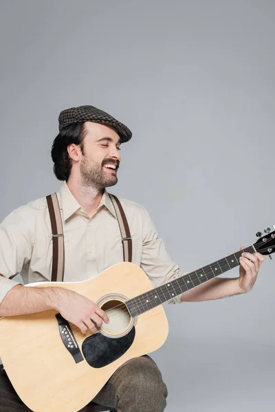 Happy man with mustache in retro style clothing and hat playing acoustic guitar isolated on grey — Stock Photo